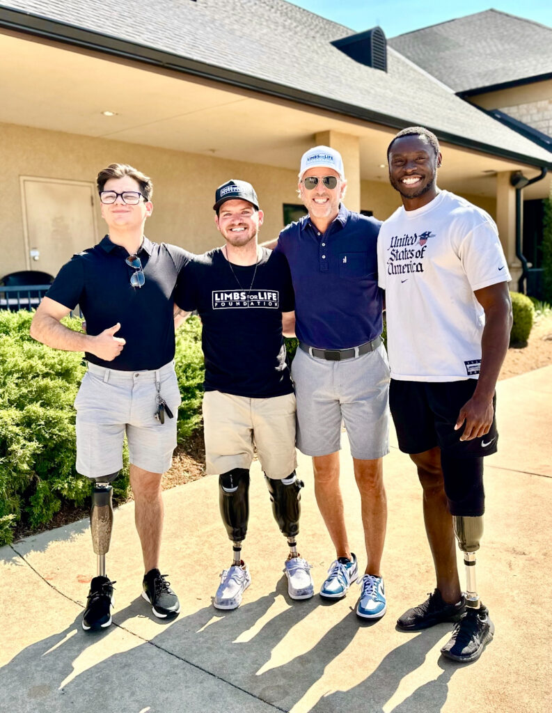Four men standing together outside a building with a shingled roof, arms around each other's shoulders, smiling at the camera. Three wear prosthetic legs. One wears a black 