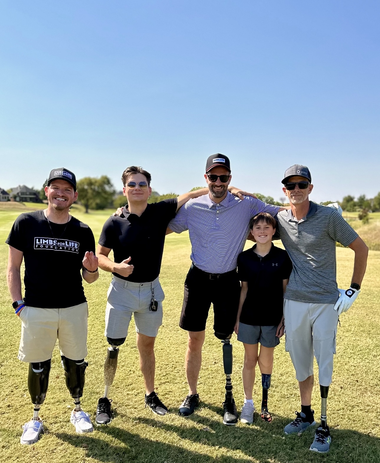 Five people posing on a golf course under a clear blue sky, arms around each other. Two men wear 
